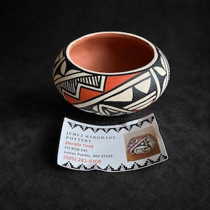 Puede incluir: Un cuenco de arcilla pequeño, pintado a mano, con patrones geométricos en negro, blanco y naranja terracota. Al lado hay una tarjeta blanca con el texto: JEMEZ HANDMADE POTTERY, Dorela Tosa, PO BOX 242, Jemez Pueblo, NM 87024, (505) 221-0709.