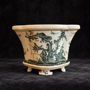 Op de afbeelding: Een crèmekleurige keramische plantenbak met een geschulpte rand en drie pootjes. De buitenkant is voorzien van een donkergroene transferprint met vissende figuren, bomen en vogels in een landschap. De basis bevat een bijpassende vaste schotel.