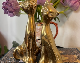 May include: Two gold-colored brass cat figurines sitting on a book with a dark blue cover and gold floral design. The cats are facing each other and have a shiny finish.