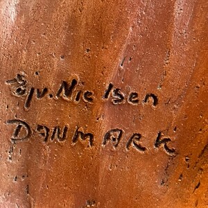 K&ouml;nnte beinhalten: Nahaufnahme einer strukturierten, rotbraunen Keramikoberfl&auml;che mit dem eingravierten Text "Sv. Nielsen Danmark". Die Oberfl&auml;che hat ein raues, organisches Aussehen mit sichtbaren Unvollkommenheiten und Farbvariationen.