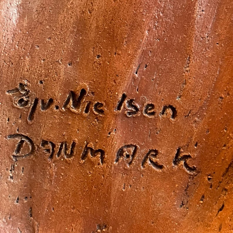 K&ouml;nnte beinhalten: Nahaufnahme einer strukturierten, rotbraunen Keramikoberfl&auml;che mit dem eingravierten Text "Sv. Nielsen Danmark". Die Oberfl&auml;che hat ein raues, organisches Aussehen mit sichtbaren Unvollkommenheiten und Farbvariationen.