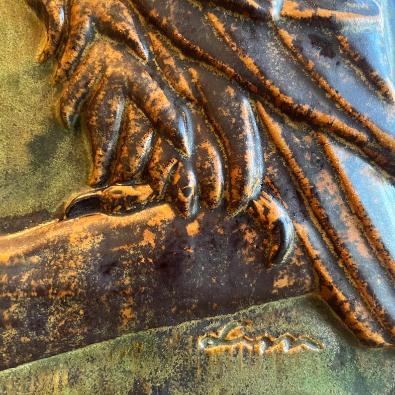 K&ouml;nnte beinhalten: Nahaufnahme einer bronzefarbenen Reliefskulptur, die eine Hand zeigt, die einen Ast oder einen Fl&uuml;gel h&auml;lt. Die strukturierte Oberfl&auml;che hat dunkelbraune und orangefarbene Farbt&ouml;ne, vor einem gr&uuml;nen Hintergrund. Die Signatur des K&uuml;nstlers ist sichtbar.