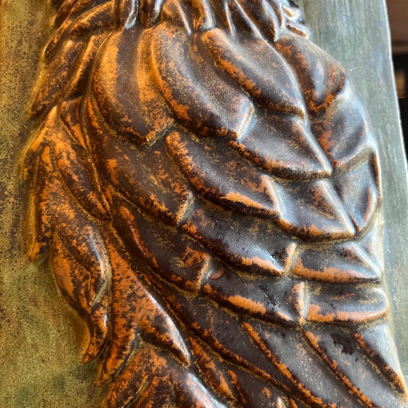 K&ouml;nnte beinhalten: Nahaufnahme einer dekorativen Eulen-Skulptur. Die Skulptur zeigt detaillierte, strukturierte Federn in Braun- und Oranget&ouml;nen mit einem verwitterten, antiken Finish. Der Hintergrund ist ein ged&auml;mpftes Gr&uuml;n, das einen Kontrast zum Gefieder der Eule bildet.