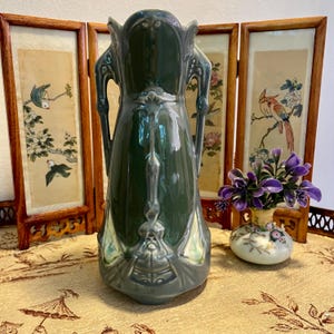 Pode incluir: Um vaso de cer&acirc;mica verde com acabamento brilhante e al&ccedil;as ornamentadas. O vaso tem um topo alargado e um design em relevo detalhado. O fundo inclui uma tela dobr&aacute;vel decorativa com pain&eacute;is pintados.