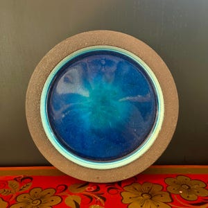 Puede incluir: Un plato redondo de cerámica con un borde marrón oscuro y un centro azul vibrante. El centro tiene un efecto degradado, que pasa de un azul profundo a un tono turquesa más claro. El diseño del plato es simple pero llamativo.