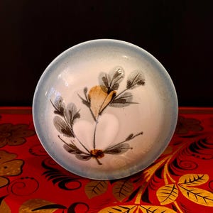 Può includere: Una piccola ciotola in ceramica bianca con un bordo blu e un motivo floreale. La ciotola presenta un fiore stilizzato con petali marroni e gialli e foglie verdi.