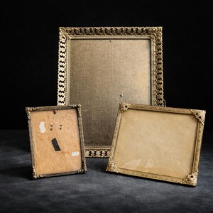 May include: Three antique gold-colored picture frames of varying sizes. The frames have ornate, detailed designs. The largest frame is rectangular, while the smaller frames are square and rectangular. The frames are empty, with visible backing.