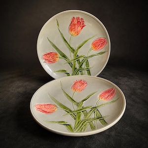 May include: Two ceramic plates with a hand-painted tulip design. The tulips are red and yellow with green stems and leaves, set against a light gray background. The plates are round and have a slightly raised edge.