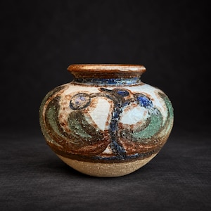 Op de afbeelding: Een kleine, ronde keramische vaas met een brede opening en een textuurrijk, gespikkeld oppervlak. De buitenkant heeft een handgeschilderd ontwerp in tinten bruin, saliegroen en diepblauw, met een crèmekleurige basis en een donkere, ruwe onderkant.