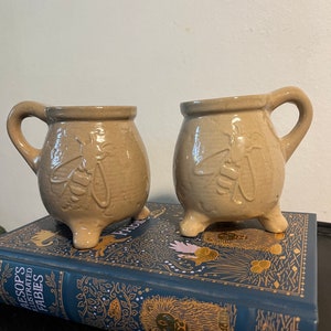 May include: Two beige ceramic mugs with bee designs and three legs each. The mugs are sitting on top of a blue book with gold illustrations.