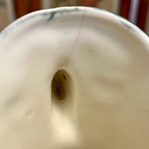Pode incluir: Close-up de um objeto de cer&acirc;mica creme com formato arredondado e um orif&iacute;cio. Uma fina rachadura percorre a lateral. A borda tem uma guarni&ccedil;&atilde;o azul-esverdeada. O interior &eacute; liso e o orif&iacute;cio &eacute; escuro.