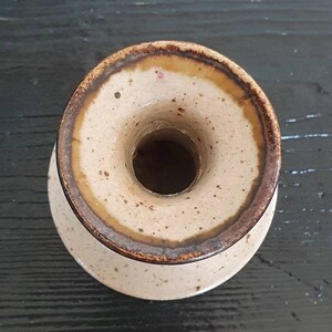 May include: A small, light brown ceramic vase with a brown rim and speckled glaze. The vase has a wide opening at the top and a narrow base.