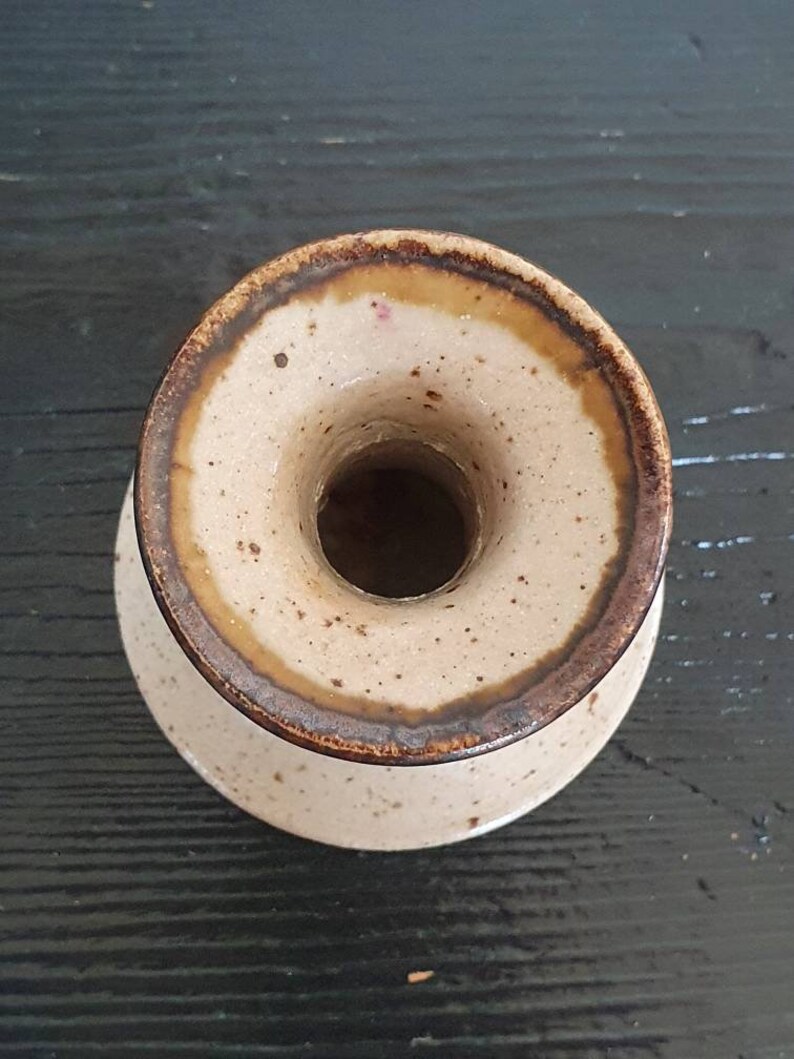 May include: A small, light brown ceramic vase with a brown rim and speckled glaze. The vase has a wide opening at the top and a narrow base.