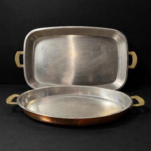 Puede incluir: Dos bandejas de servicio de metal. La bandeja superior es rectangular con asas de latón. La bandeja inferior es redonda con base color cobre y asas de latón. Ambas bandejas tienen un acabado de metal cepillado sobre un fondo negro.