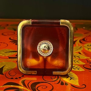 Op de afbeelding: Een kleine, vierkante, rode, doorzichtige plastic doos met een gouden rand en een gouden ronde knop aan de bovenkant.