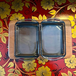 Puede incluir: Dos fuentes para hornear de vidrio transparente con asas sobre una tabla de cortar de madera. La tabla de cortar está sobre una superficie roja con un patrón floral.