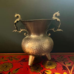 Puede incluir: Un jarrón de metal martillado con tres patas y asas ornamentadas con figuras de dragones. El jarrón tiene una boca ancha y un cuerpo bulboso. Las asas están detalladas con diseños arremolinados. El jarrón es probablemente de latón o bronce.