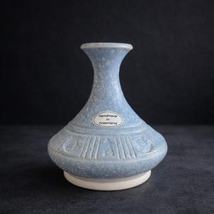 May include: A pale blue ceramic vase with a speckled finish and a broad base. The vase features a slender neck and a decorative band with carved patterns. A sticker on the vase reads "Handmade in Greenland". It rests on a white base.