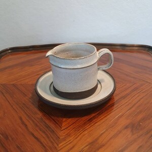 May include: A small white ceramic creamer with a black rim and a matching saucer. The creamer has a handle and a spout.
