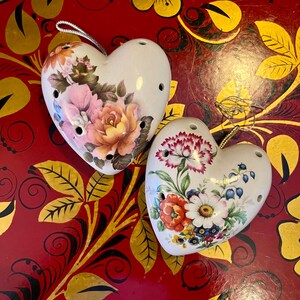 Può includere: Due ornamenti in ceramica bianca a forma di cuore con motivi floreali. I cuori sono decorati con fiori rosa, gialli e blu e foglie verdi. I cuori sono su uno sfondo rosso con motivi floreali dorati e neri.