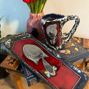 Könnte beinhalten: Ein schwarzes und rotes Keramiktablett mit einem stilisierten Frauendesign. Das Tablett ist mit goldenen Akzenten verziert und hat eine strukturierte Oberfläche. Eine schwarz-weiße Keramiktasse mit einem stilisierten Frauendesign befindet sich im Hintergrund.