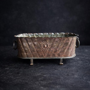 Pode incluir: Uma floreira retangular de cobre com um exterior texturizado com padrão de diamante. Possui pequenas alças curvas em cada lado e é suportada por quatro pés pequenos. Um design circular decorativo está centrado na frente.