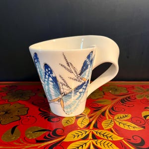 May include: White ceramic mug with a unique handle design. The mug features a blue and white butterfly design with delicate floral accents. The mug is set against a red and gold decorative surface.