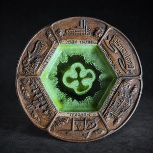 May include: A round brown ceramic souvenir dish featuring a central hexagonal green glazed well. The rim displays embossed relief carvings of Disneyland attractions including the Mark Twain, Jungle Cruise, Sleeping Beauty Castle, Monorail, and Haunted Mansion.