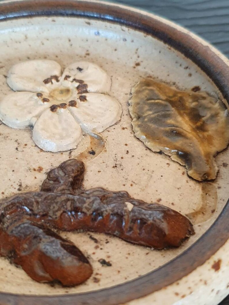May include: A ceramic dish with a brown rim and a white interior. The dish features a brown, leaf-shaped design, a white flower with a brown center, and a brown, abstract design.
