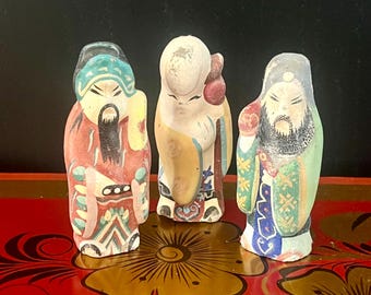 以下が含まれることがあります： それぞれ異なるカラフルなローブと表情を持つ、中国の神々の小さなセラミック製フィギュア3体。フィギュアは、金色の花柄が施された赤い表面の上に立っています。