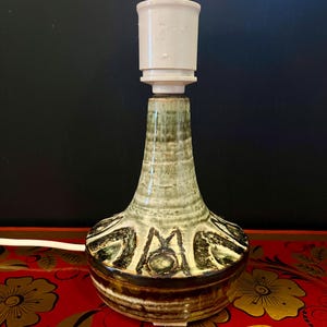Könnte beinhalten: Eine Keramik-Tischlampe mit grüner und brauner Glasur. Die Lampe hat einen weißen Keramikschirm und ein dekoratives Muster um die Basis.