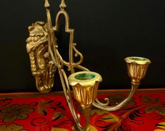May include: A gold-toned metal wall sconce with a decorative dragon head and two candle holders. The sconce is on a red and gold patterned surface.