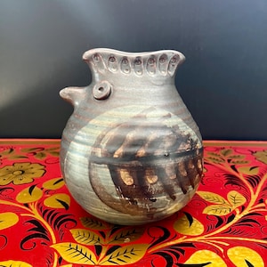 Puede incluir: Un jarrón de cerámica marrón y gris con forma de pájaro, con una superficie texturizada y un diseño marrón y blanco. El jarrón tiene un cuerpo redondo y un pequeño pico.