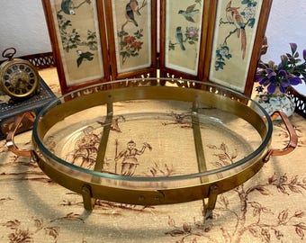 May include: An oval glass serving dish with a brass-colored metal frame and copper handles. The dish is supported by four small legs. The background includes a folding screen with bird and floral artwork.