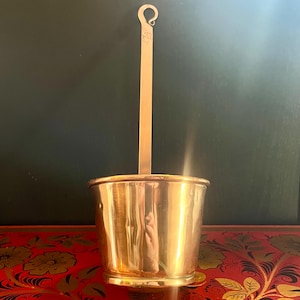 May include: A vintage brass bucket with a long handle and a looped end. The bucket has a cylindrical shape and a slightly flared base. The handle is attached to the center of the bucket and extends upwards. The brass has a warm, golden hue.