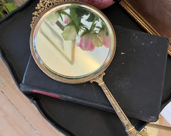 May include: A gold-toned hand mirror with an ornate frame and handle. The mirror is reflecting a pink and white flower.