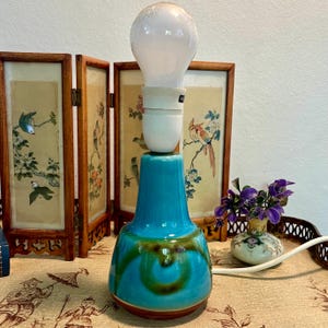 May include: A vintage ceramic table lamp with a turquoise base and a large light bulb. The base features a brown bottom and green and brown swirled accents. The lamp is placed in front of a decorative folding screen with bird illustrations.