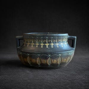 May include: A matte black ceramic bowl with two side handles, featuring a decorative band of embossed circles and gold-toned accents. The vessel has a wide, rounded body and a textured geometric pattern encircling the upper and lower sections.