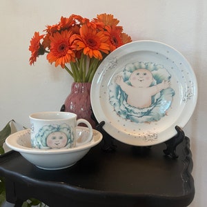 Könnte beinhalten: Ein weißes Kindergeschirr-Set aus Keramik mit einem Cabbage Patch Kid-Design. Das Set enthält eine Schüssel, einen Teller und eine Tasse. Schüssel und Tasse zeigen ein Cabbage Patch Kid-Gesicht mit einem grünen Kohlblatt um den Kopf. Der Teller zeigt ein Cabbage Patch Kid, das in einem grünen Kohlblatt liegt.