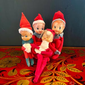May include: Four vintage Christmas elf dolls. The dolls have red outfits and hats with white trim. One elf is lying down, appearing to be asleep. The dolls are on a red surface with gold floral designs.