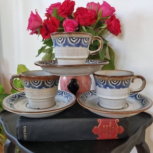 Könnte beinhalten: Drei Keramik-Kaffeetassen mit braunen und blauen Mustern und passenden Untertassen. Die Tassen und Untertassen stehen auf einem schwarzen Tisch mit einem Buch.