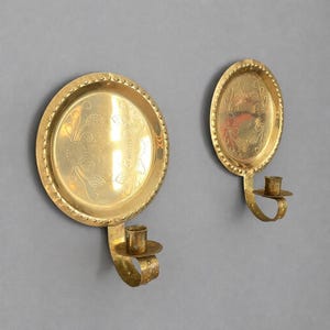 May include: Two antique brass wall sconces. One is round, the other oval, both with ornate floral designs and a small candle holder. The sconces are mounted on a gray wall, showcasing their warm, golden hue.