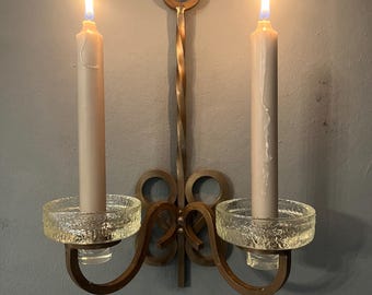 Peut inclure: Un bougeoir mural en laiton avec deux bougies allumées. Le bougeoir a un design en métal torsadé et des coupelles à bougies en verre transparent.