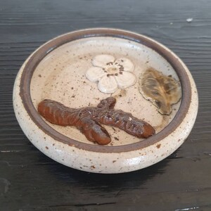 May include: A small, round, ceramic dish with a speckled glaze. The dish features a brown twig design with a white flower and a green leaf.
