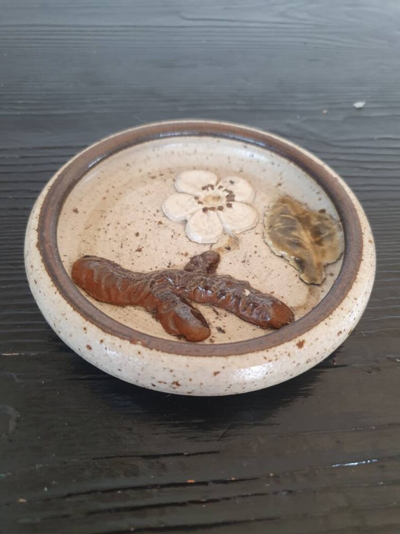May include: A small, round, ceramic dish with a speckled glaze. The dish features a brown twig design with a white flower and a green leaf.