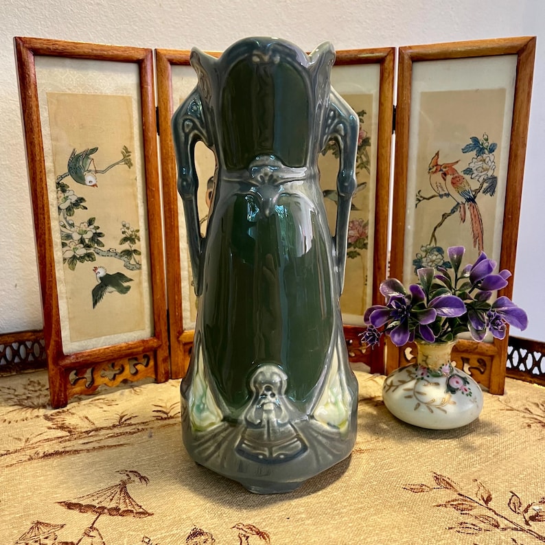 Pode incluir: Um vaso de cer&acirc;mica verde escuro com acabamento brilhante, al&ccedil;as ornamentadas e base detalhada. O vaso &eacute; exibido em frente a um biombo com obras de arte de inspira&ccedil;&atilde;o asi&aacute;tica. H&aacute; tamb&eacute;m um pequeno vaso com flores roxas.