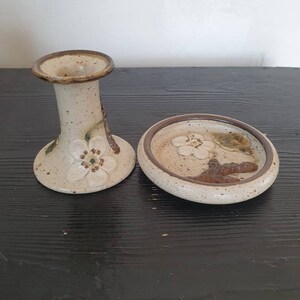 May include: A set of two ceramic candle holders with a white glaze and brown accents. The holders feature a floral design with white flowers and brown leaves. The larger holder is a candlestick with a wide base and a narrow top. The smaller holder is a round dish with a raised rim.
