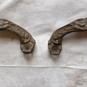 May include: Two antique bronze-toned drawer pulls with ornate floral and scrollwork designs. The curved handles have a detailed, aged appearance, suggesting vintage or repurposed hardware. They are set against a white, textured background.