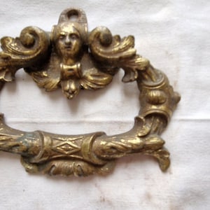 May include: Ornate antique brass drawer pull with a central female face and scrolling foliate design. The handle is a loop shape with decorative elements and a tarnished finish, suggesting age.