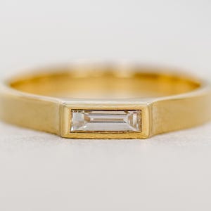 May include: A gold ring with a rectangular, clear gemstone. The ring has a simple, modern design with a brushed finish. The gemstone is set in a bezel setting, and the band is flat and wide.
